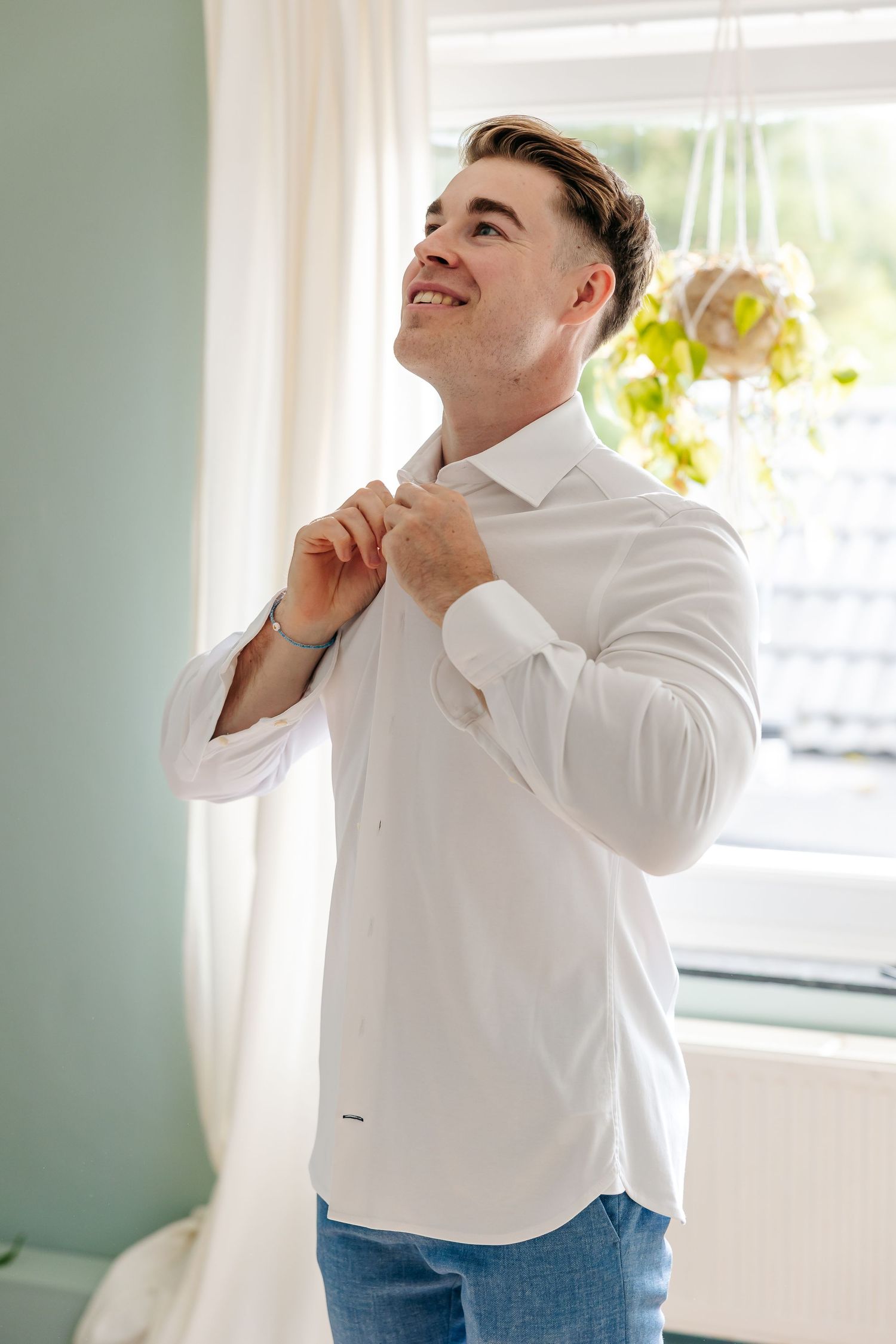 Smiling groom buttoning white dress shirt while getting ready for wedding.