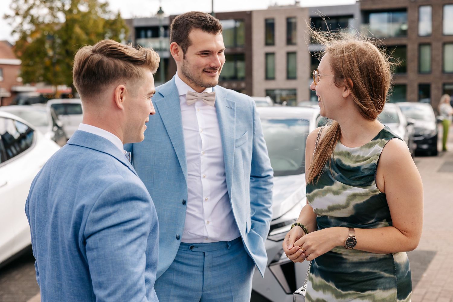 Two men in light blue suits chat with a woman in a camo dress near parked cars.