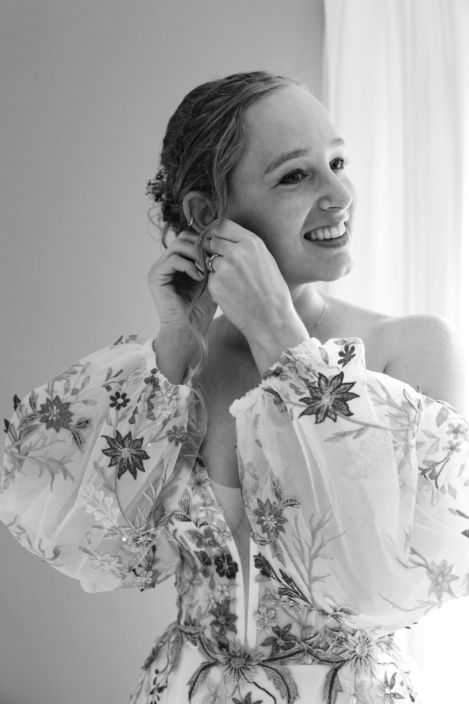 Smiling bride in floral off-shoulder gown putting on earrings, elegant black and white portrait.