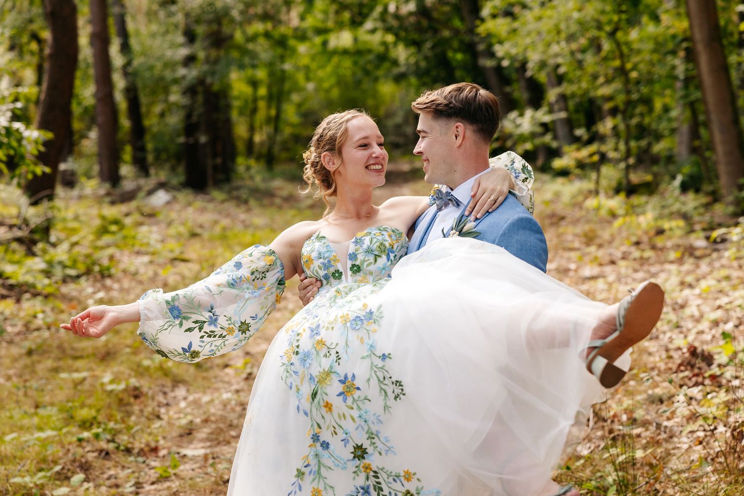 Groom carrying bride in floral wedding dress through sunlit forest path.