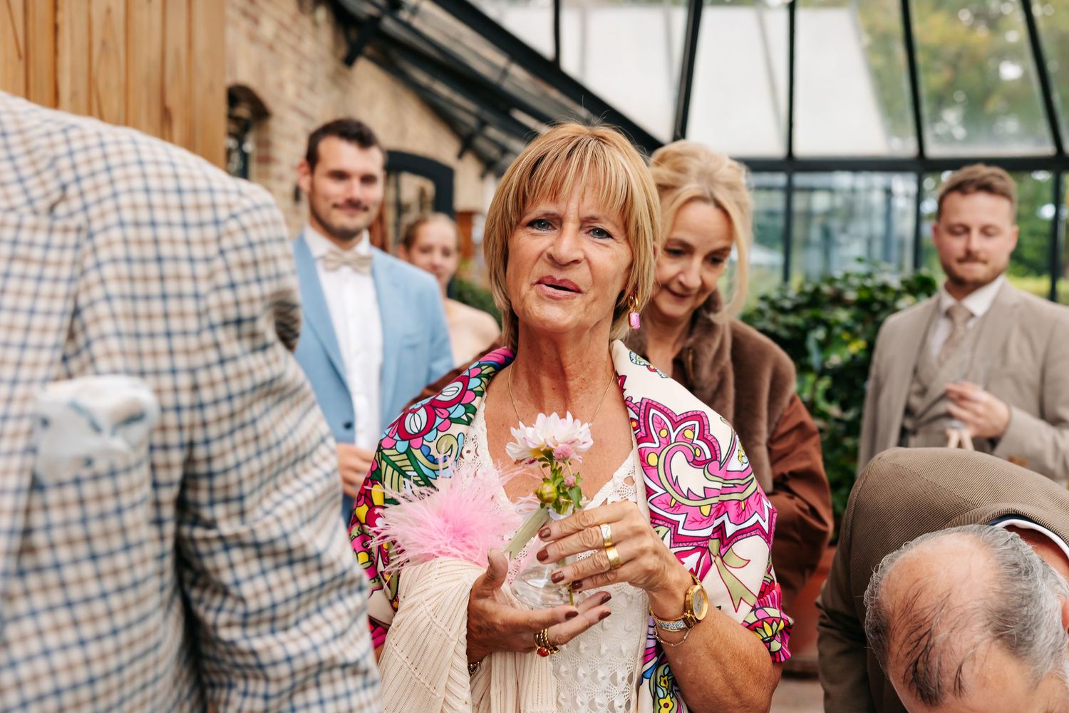 Older woman in colorful jacket holding pink flower at elegant outdoor wedding event.