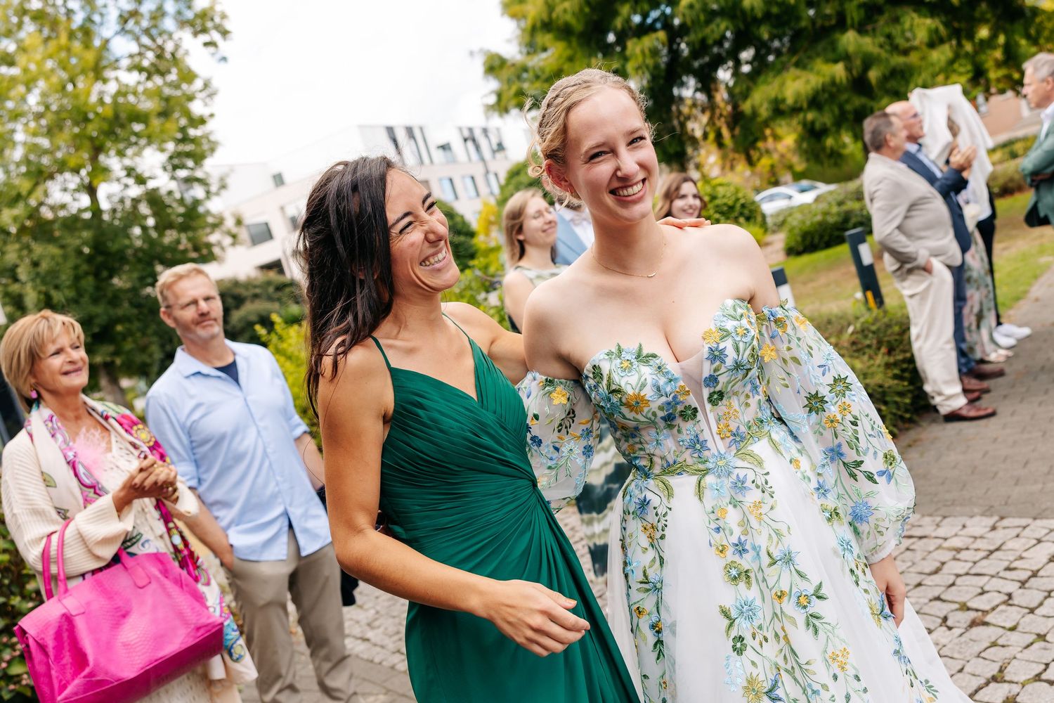 Two elegantly dressed women laugh together at an outdoor wedding event, one wearing green and one in floral.