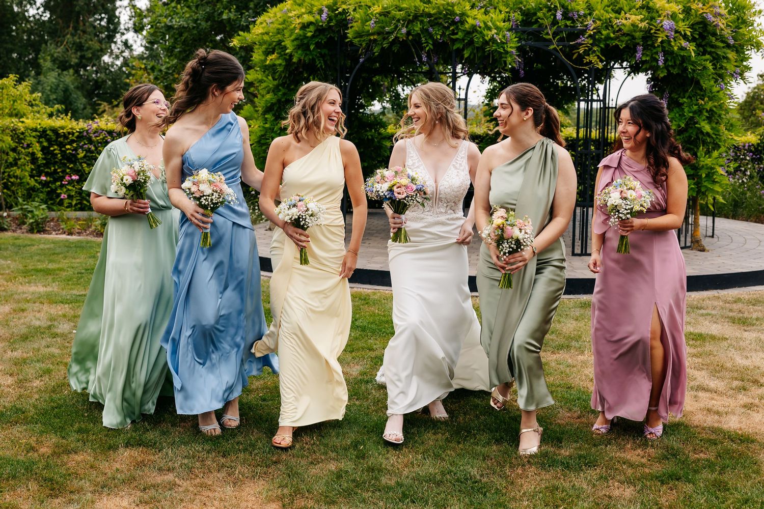 Bride and five bridesmaids in pastel mismatched dresses walk and laugh together across green garden lawn.