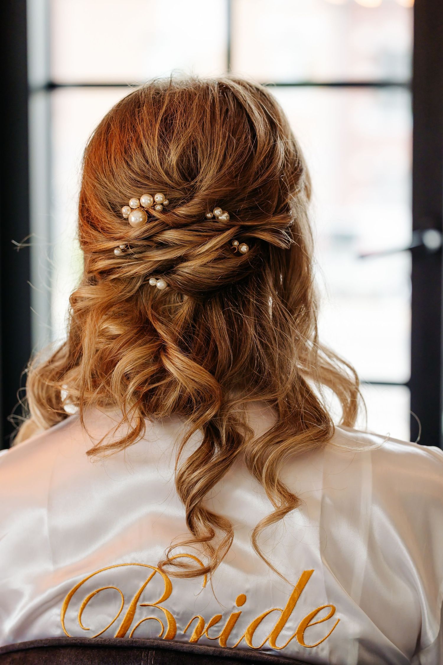 Close-up of bride's elegant half-up hairstyle with pearl hair pins, wearing white satin robe.