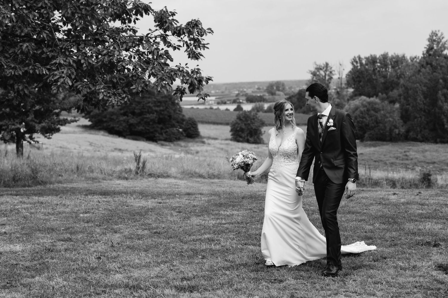 Black and white photo of a bride and groom walking together in a scenic countryside.