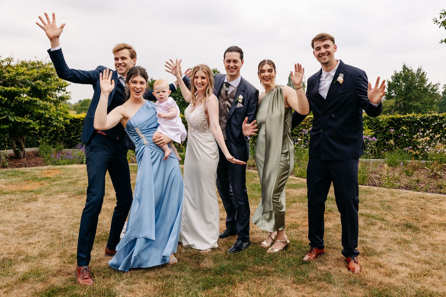 A joyful wedding party of seven poses outdoors, including bride in white and guests in colorful formal attire.