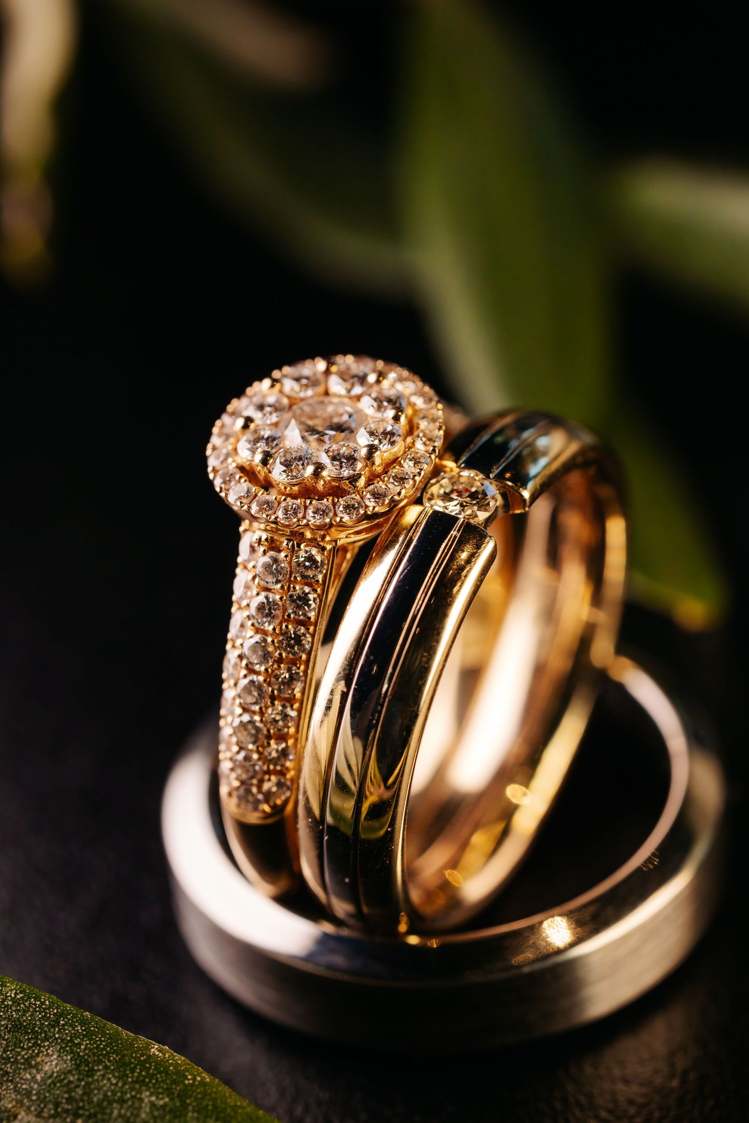 Elegant gold diamond halo engagement ring stacked with wedding bands on dark background with greenery.