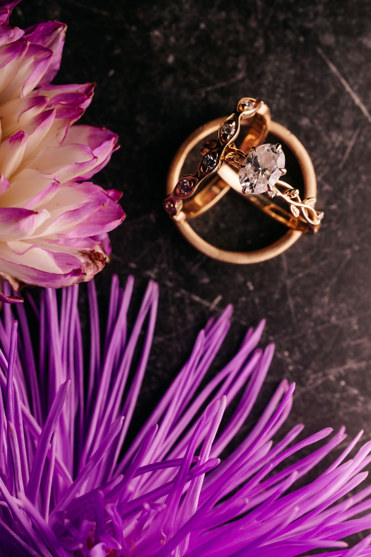 Gold engagement ring with diamond and wedding band displayed among purple and white flowers.