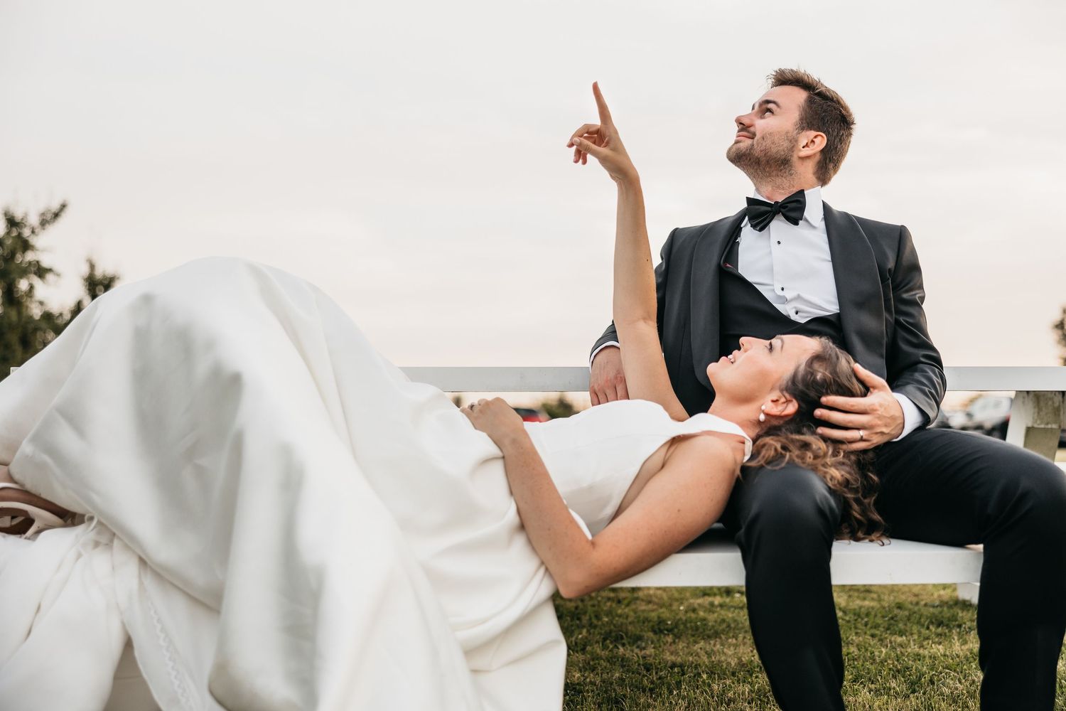 Groom in tuxedo points skyward while bride reclines across his lap on a white bench outdoors.