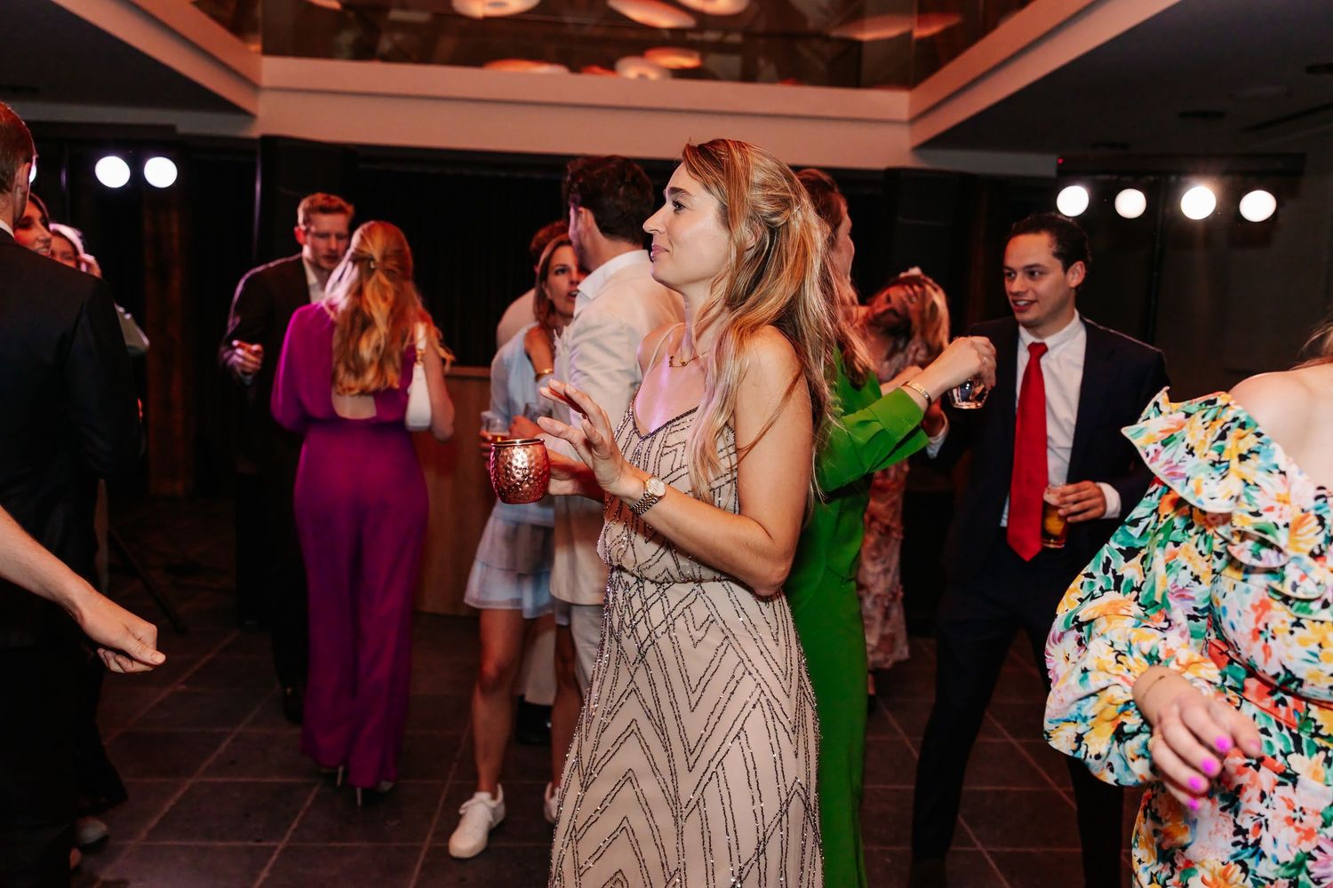 Wedding reception guests dancing and celebrating under bright lights, woman in floral dress holding drink.