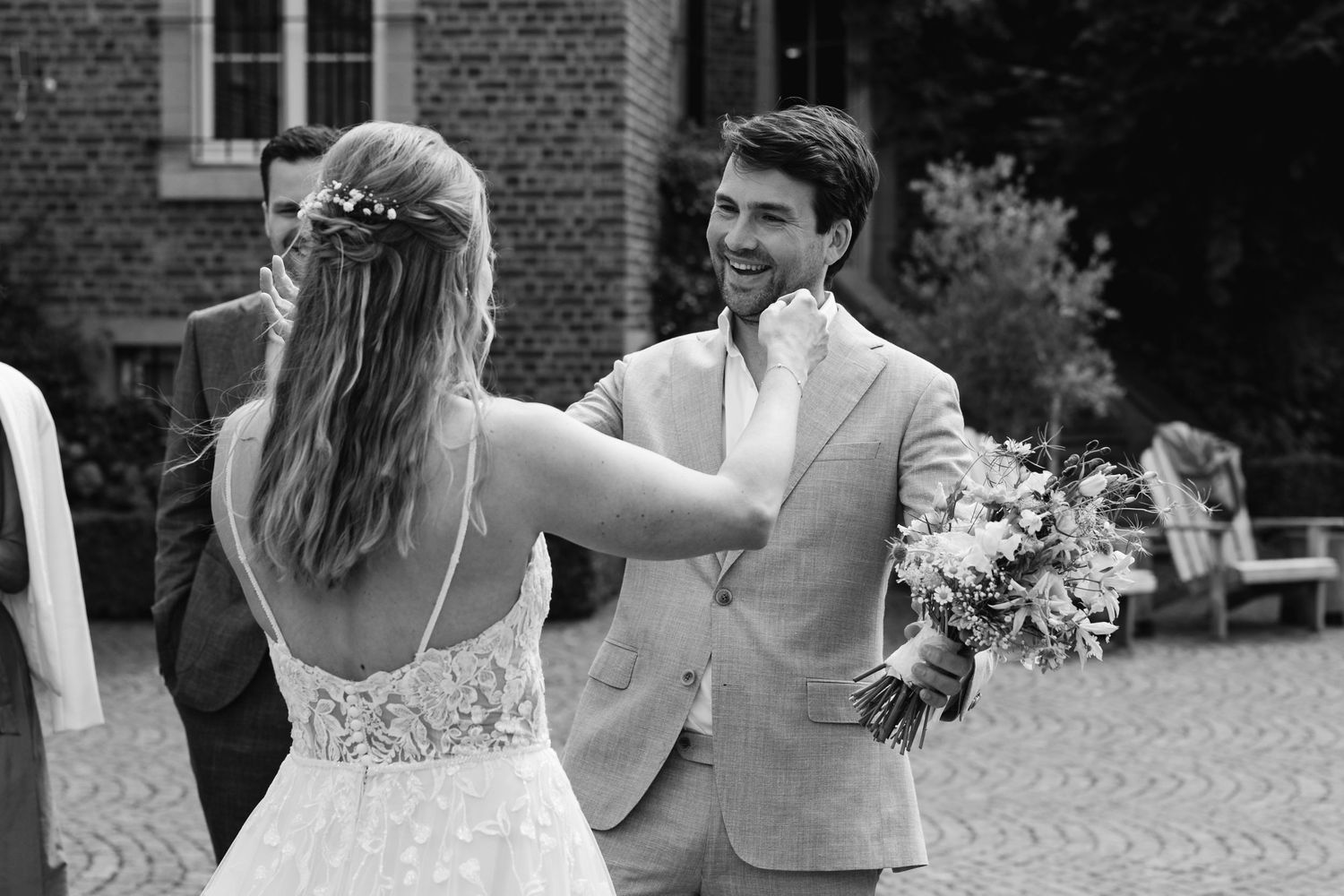 Groom in beige suit smiling at bride holding bouquet during outdoor first look ceremony.