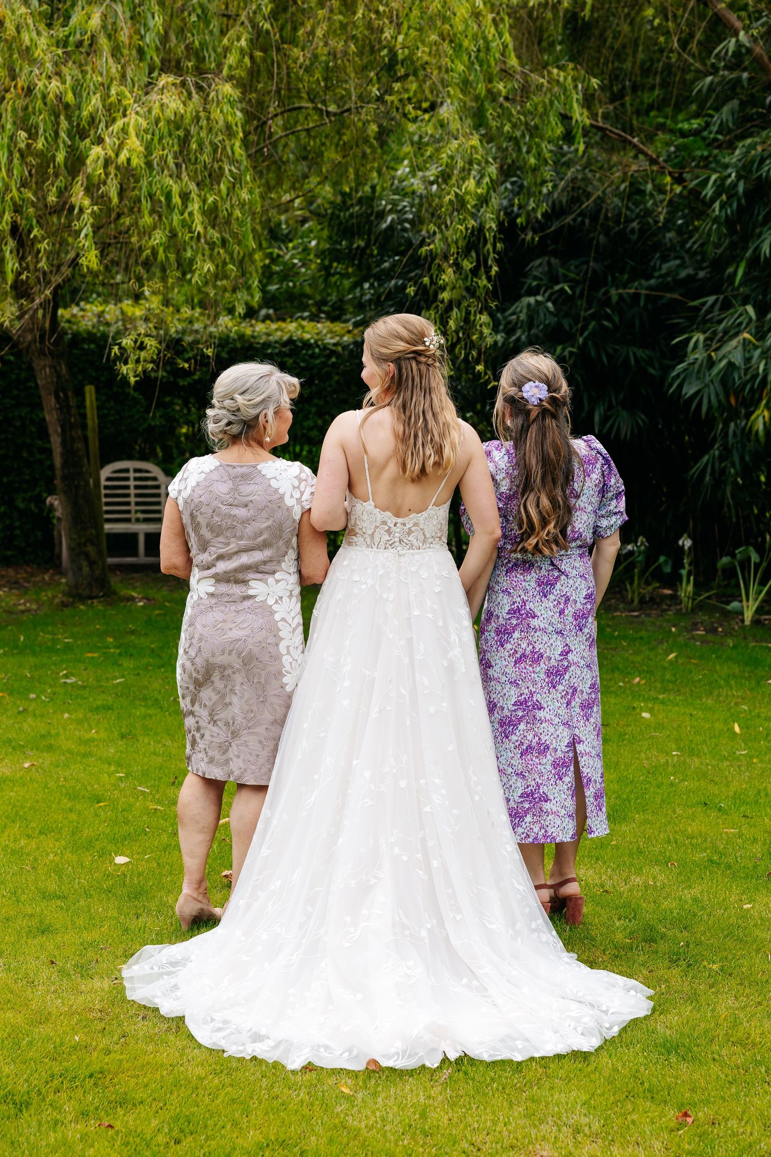 Back view of bride in lace wedding dress with two guests in lush green garden.