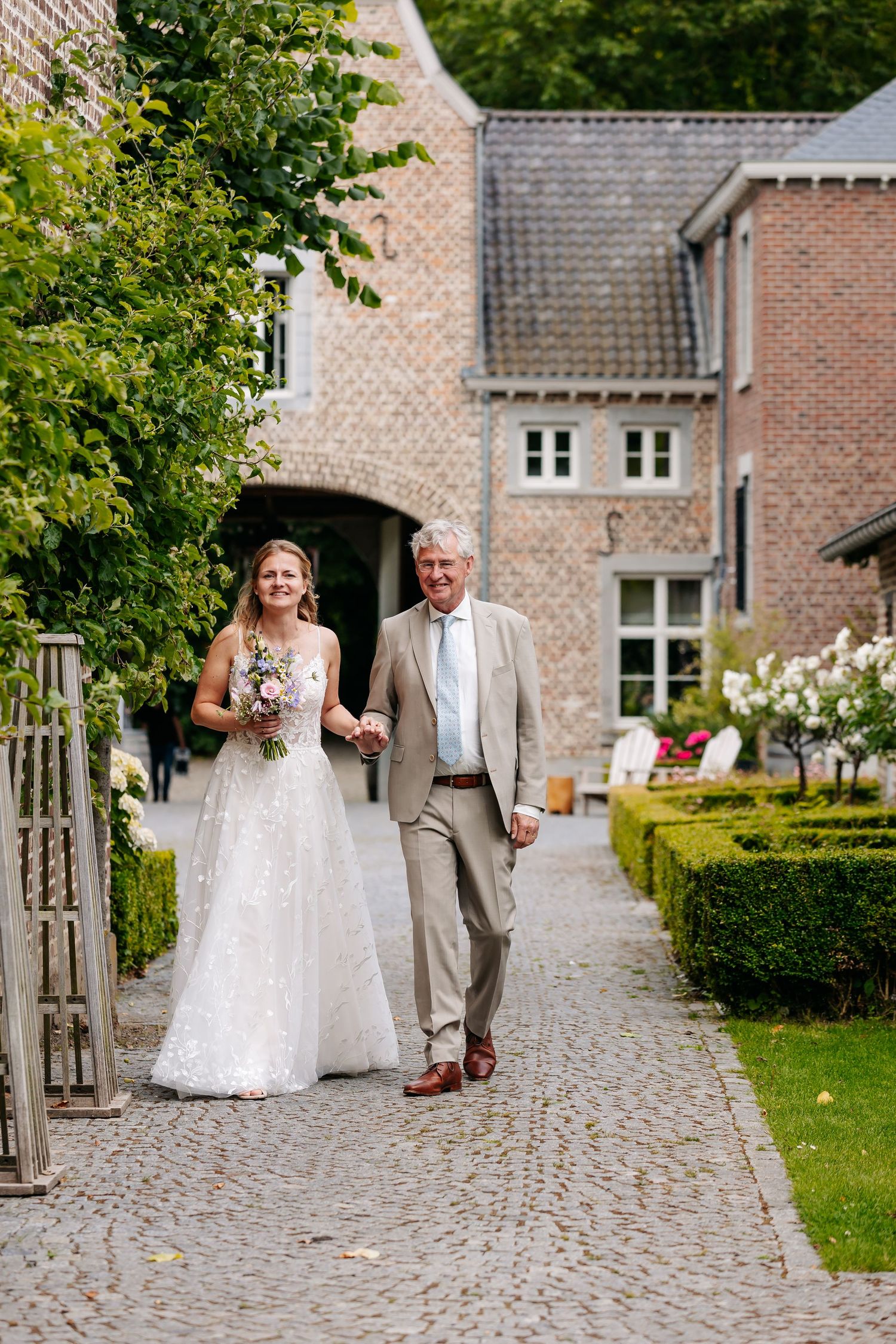 Bride in lace gown walking with older gentleman along cobblestone path beside brick wedding venue.