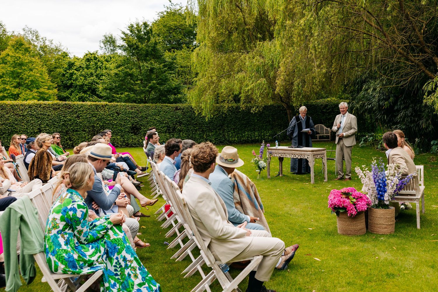 Guests seated in white chairs watch outdoor garden wedding ceremony with officiant at decorated table.