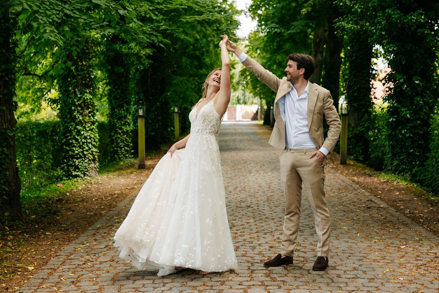 Bride and groom dancing joyfully on cobblestone path lined with tall green trees.