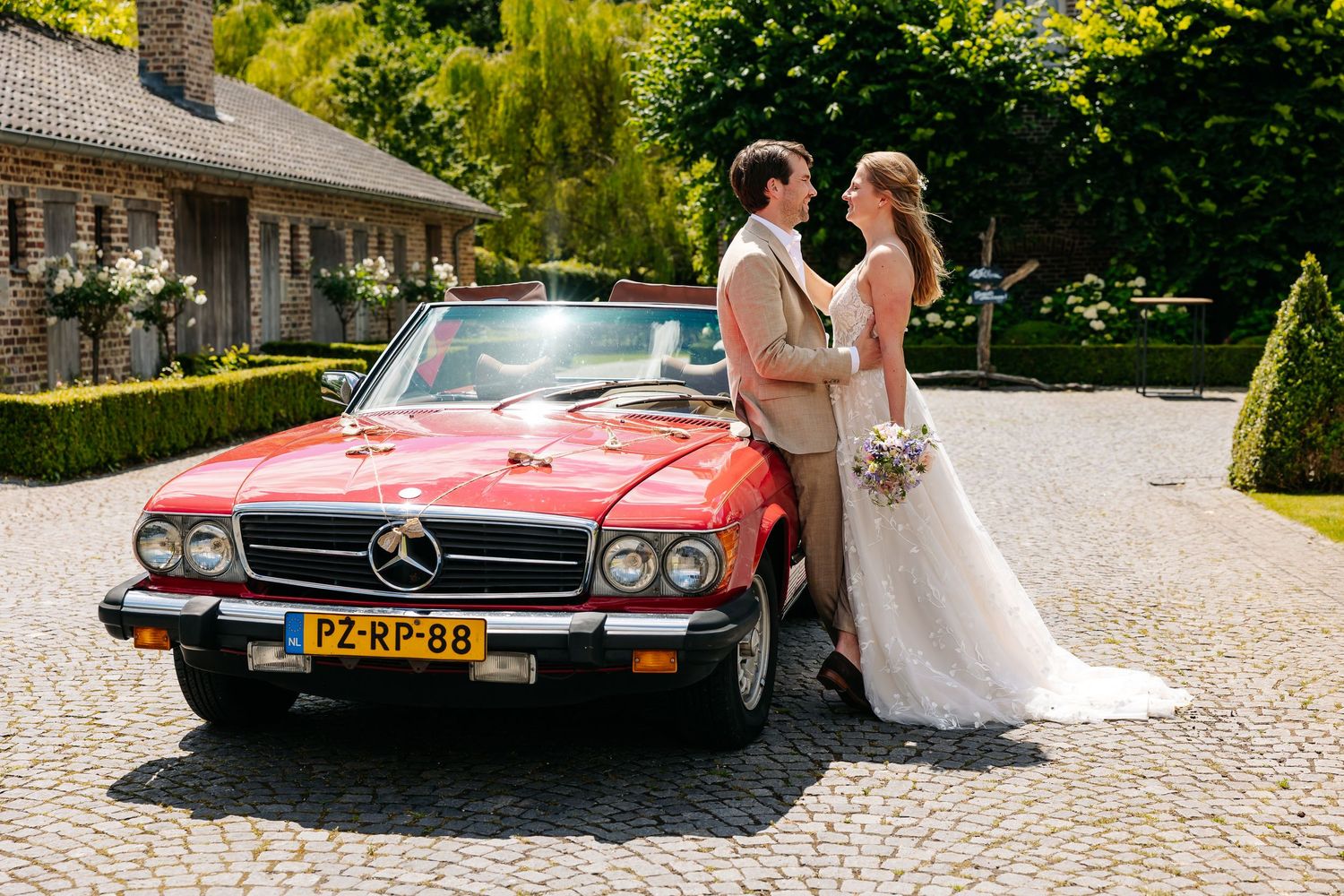 Bride and groom posing beside a classic red Mercedes convertible on wedding day.