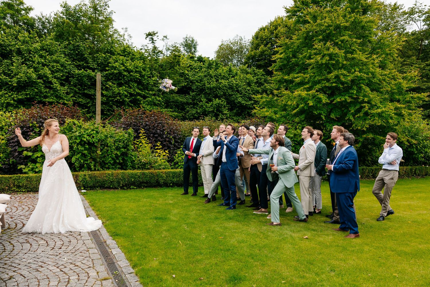 A bride in a white gown tosses her bouquet to a group of male wedding guests outdoors.