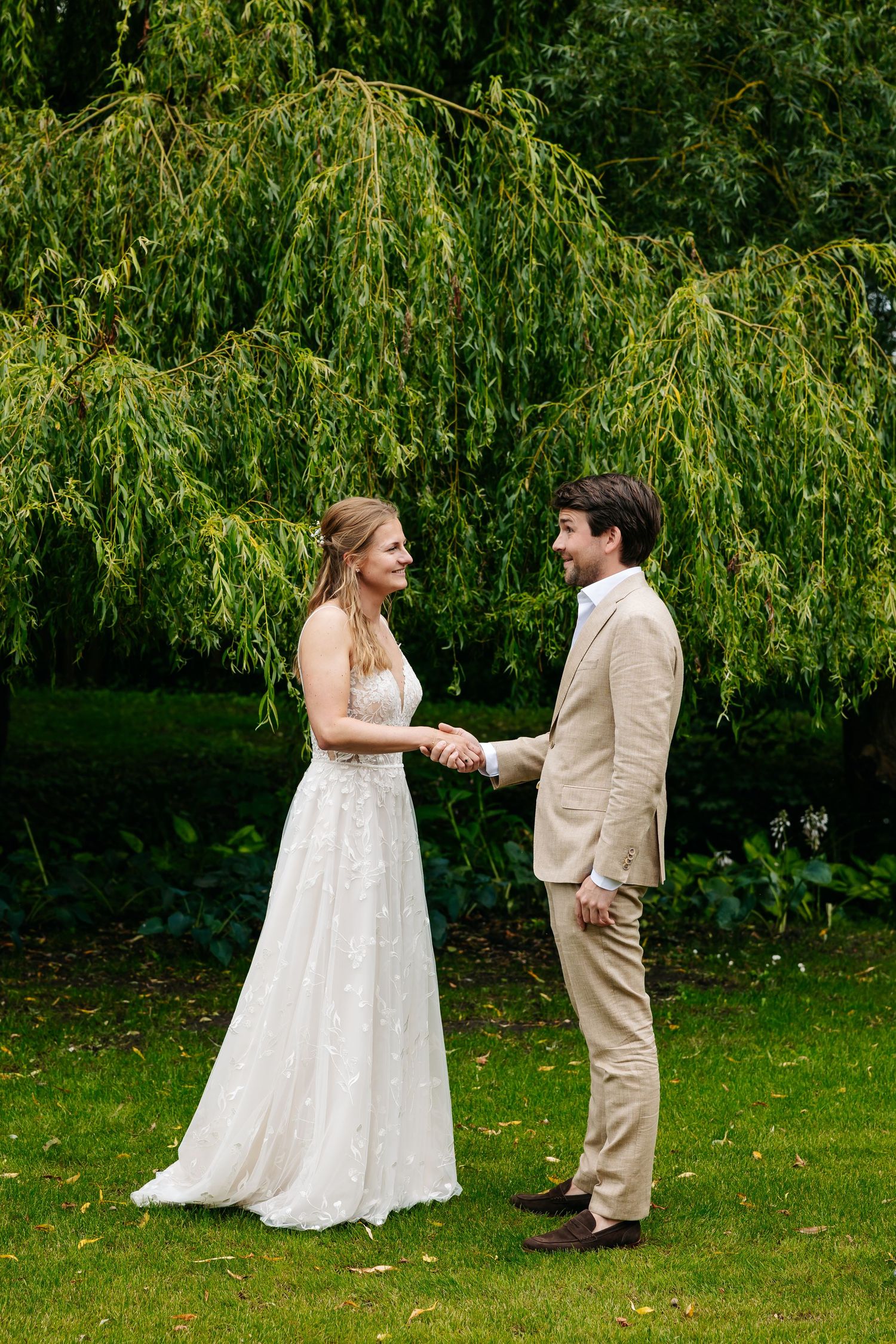 Bride and groom hold hands smiling at each other in a lush green garden during their wedding ceremony.