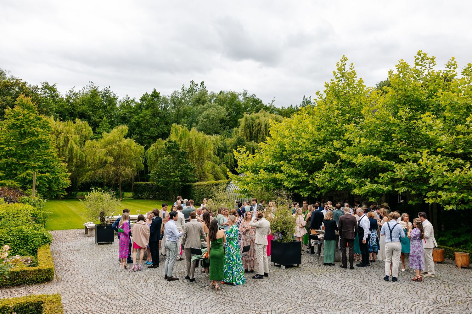 Wedding guests mingle on cobblestone terrace surrounded by lush green trees and gardens.
