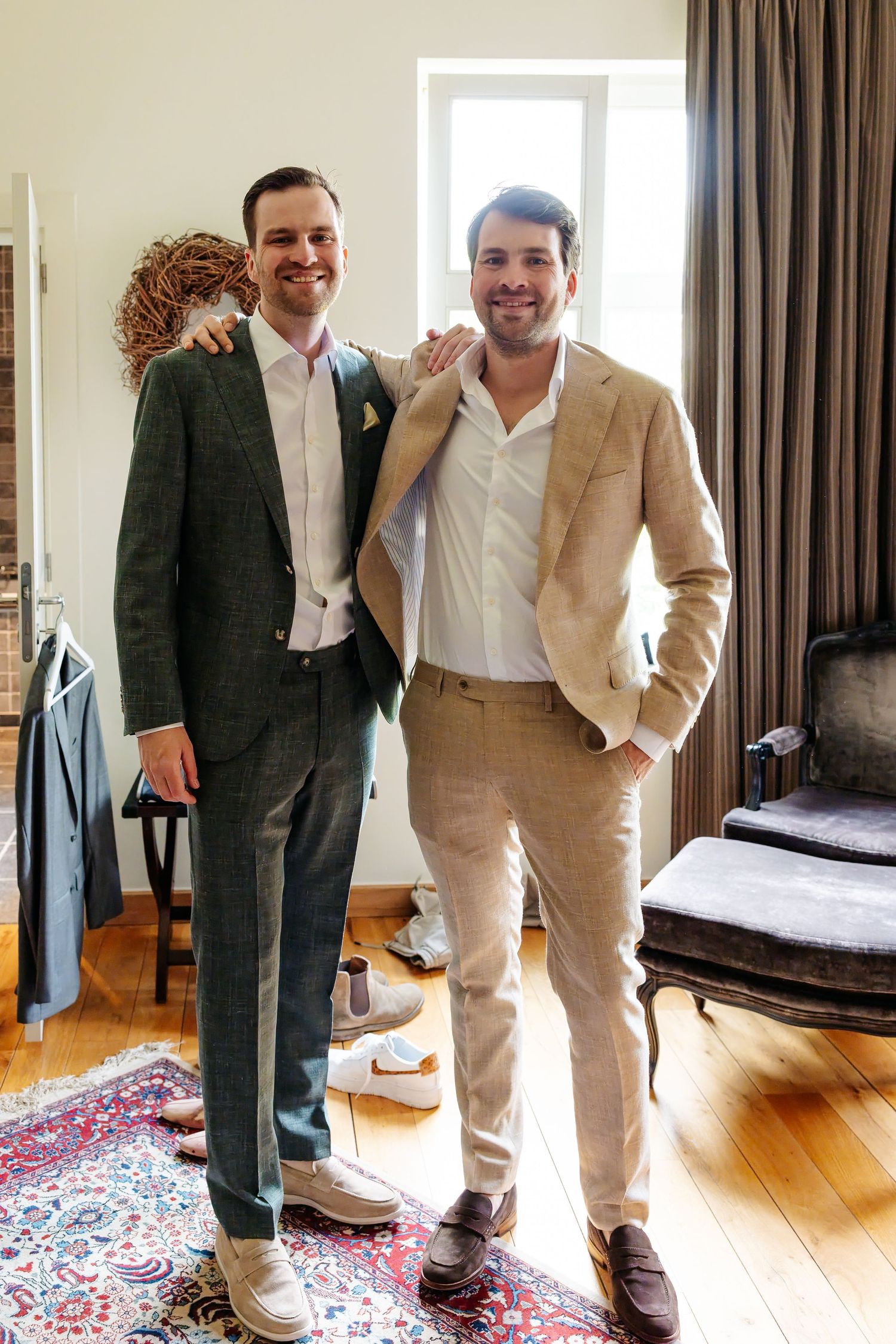 Two smiling men in suits posing together indoors on a patterned rug in a warmly lit room.