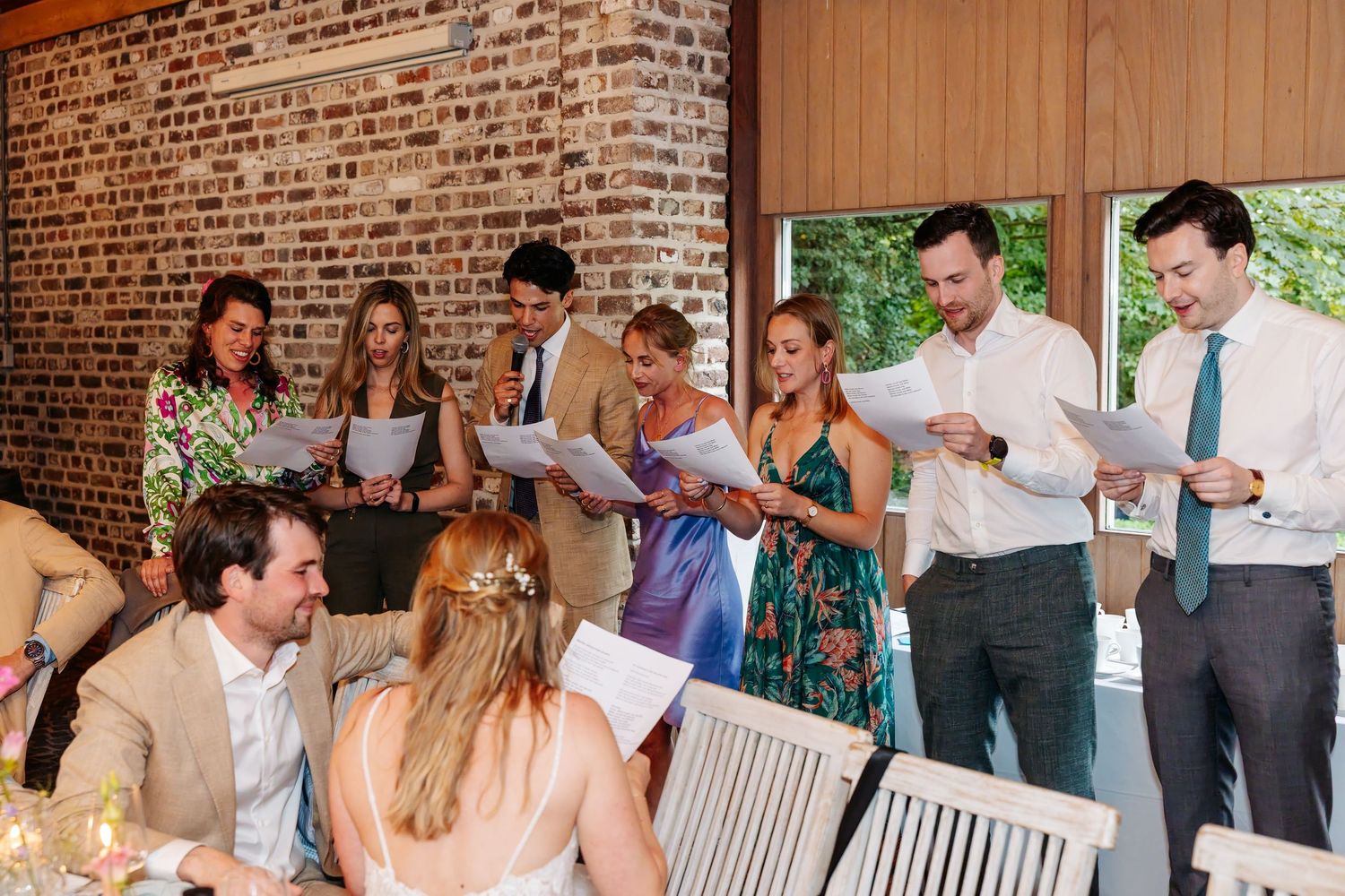 Wedding guests singing together from printed sheets at reception in brick venue.