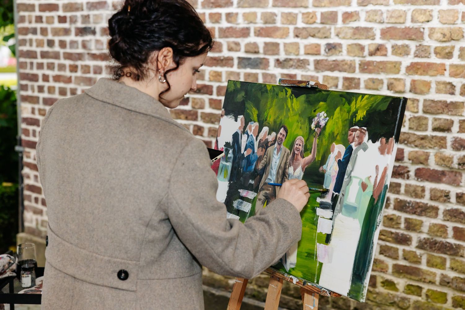 Artist painting a live wedding scene on canvas at an easel against a rustic brick wall backdrop.