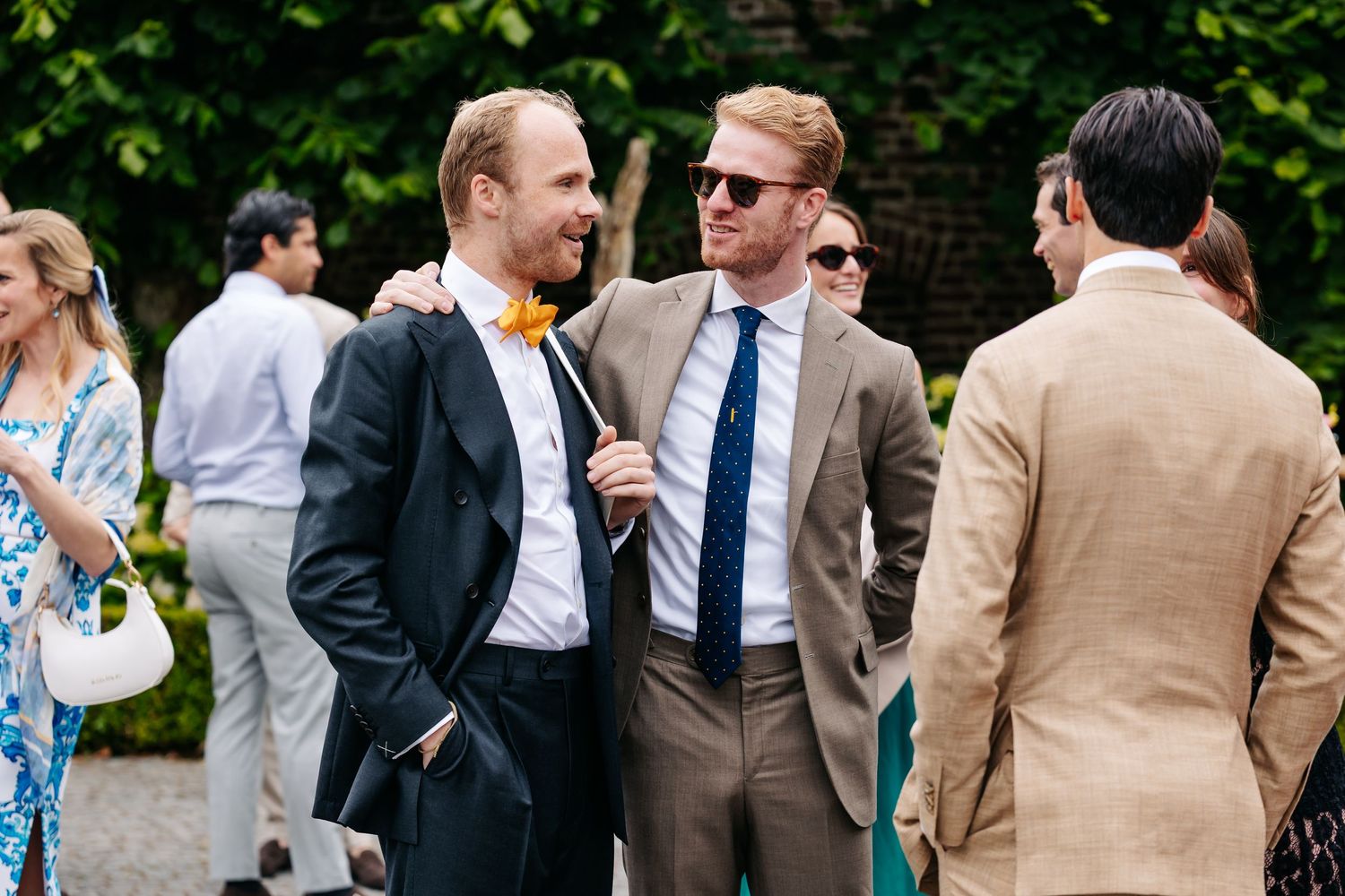 Two well-dressed men in suits share a laugh at an outdoor wedding reception in a garden setting.