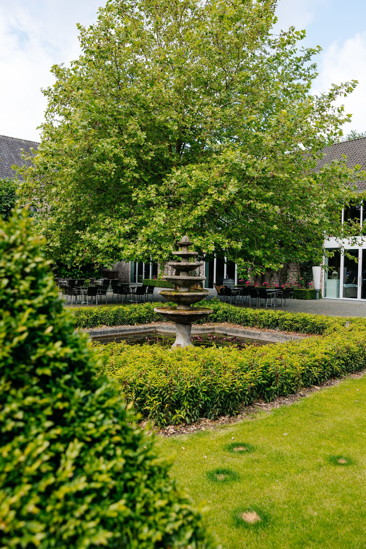 Elegant garden with tiered stone fountain, manicured hedges, and a large green tree outside a brick building.