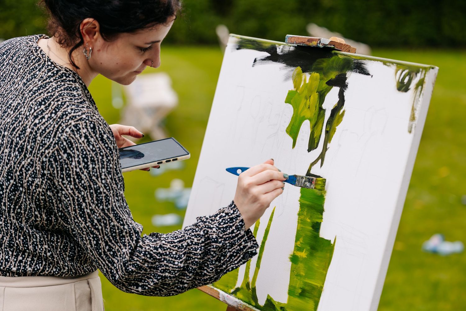 Woman painting on a canvas at an easel outdoors on a green lawn during a wedding event.