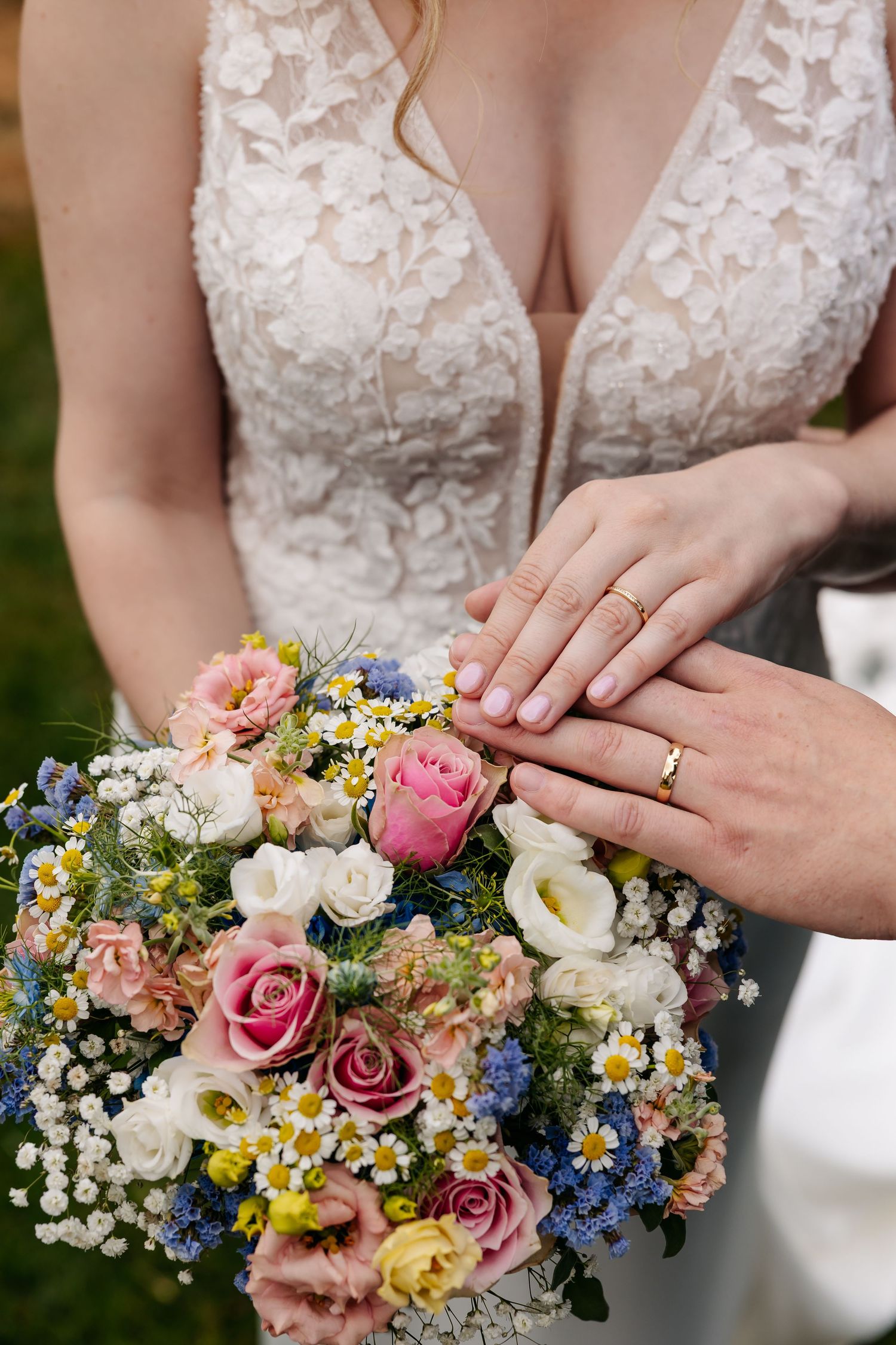 Newlyweds displaying gold wedding rings over colorful bridal bouquet with roses and wildflowers.