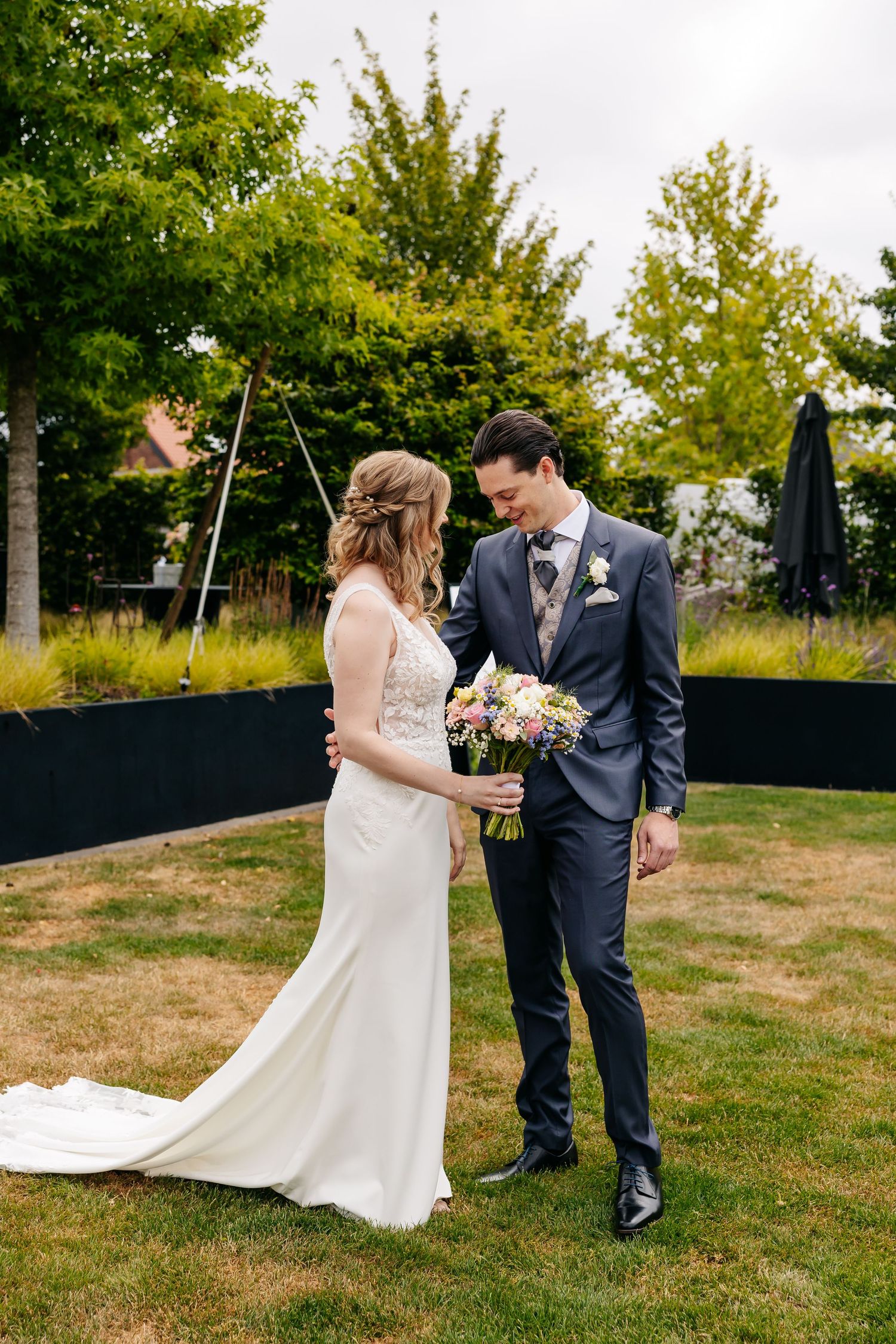 Bride and groom share a tender moment on garden lawn, admiring bridal bouquet of pink and white flowers.