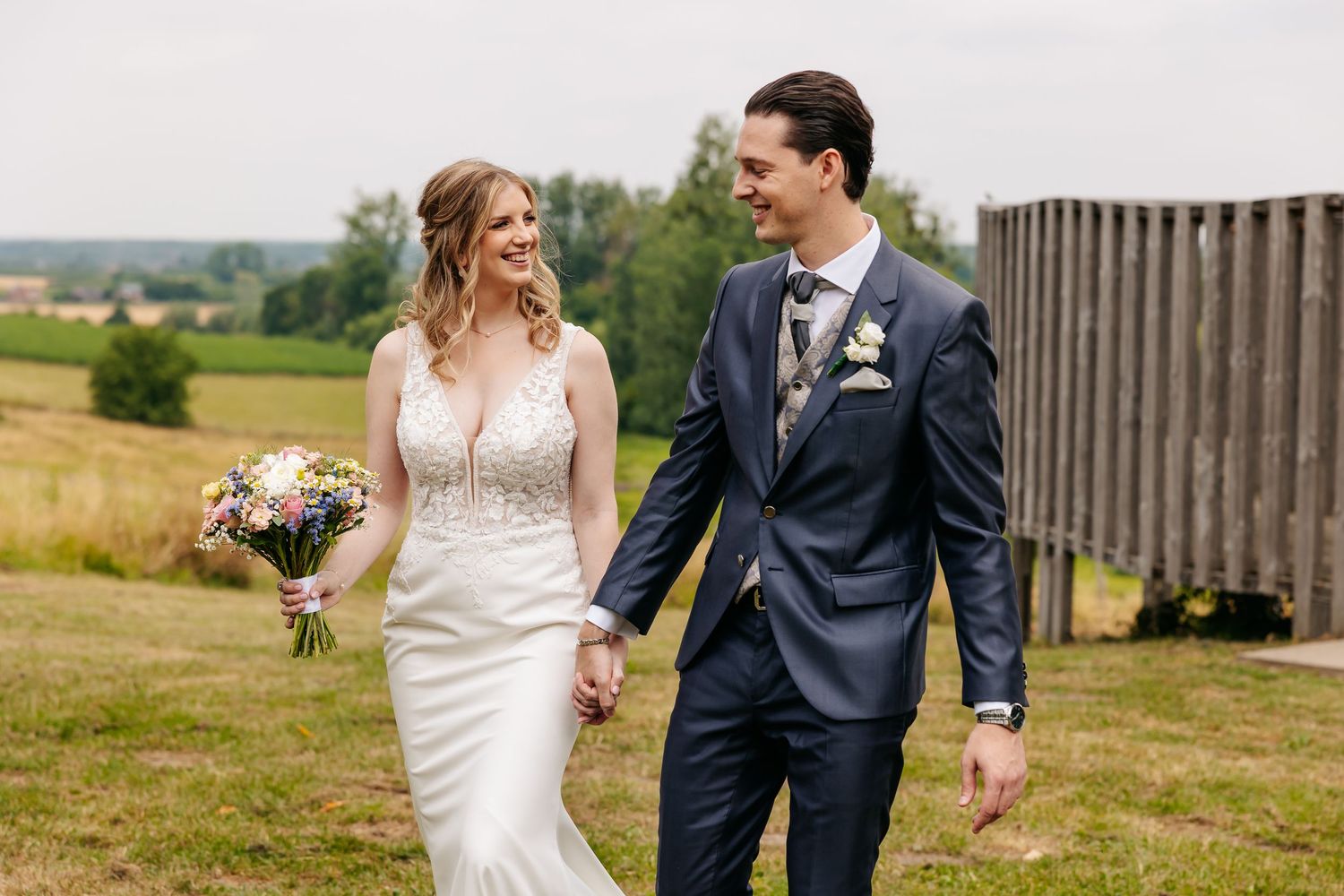 Laughing bride and groom holding hands while strolling through a sunny countryside field on wedding day.