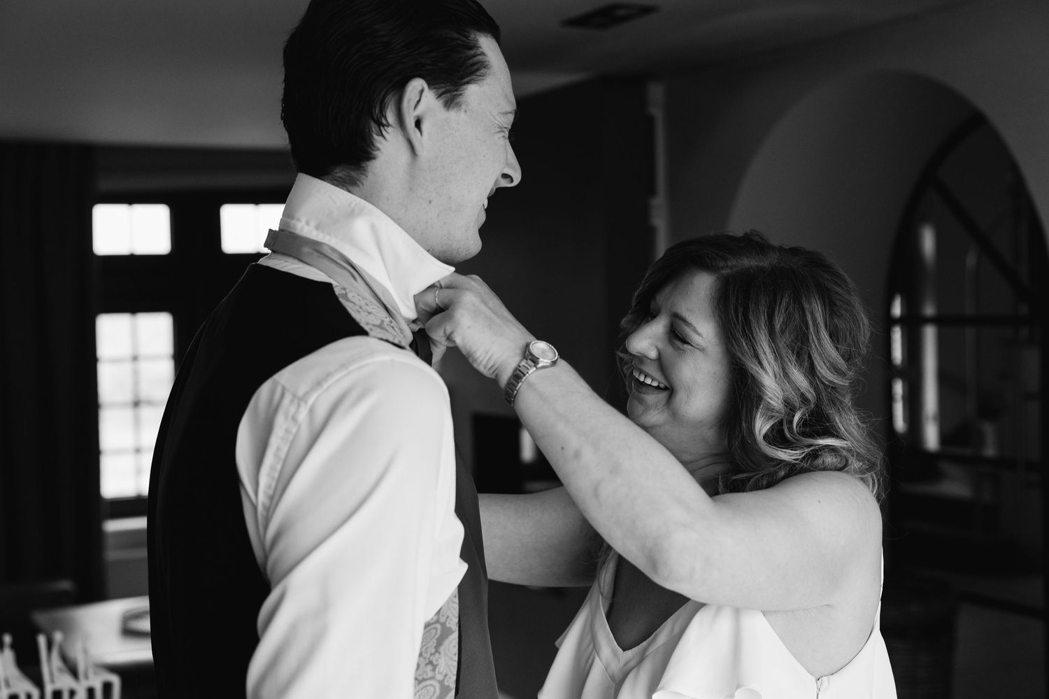 Black and white photo of woman helping groom adjust his tie while smiling warmly.