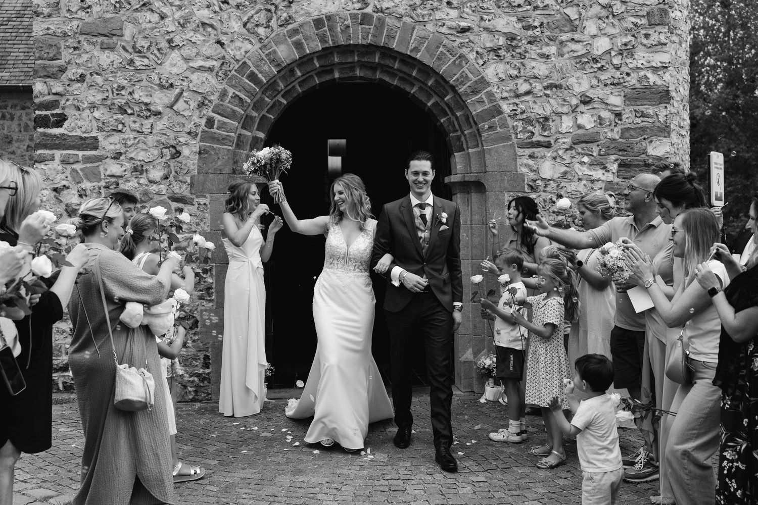 Black and white photo of smiling couple exiting rustic stone church arch surrounded by cheering guests.