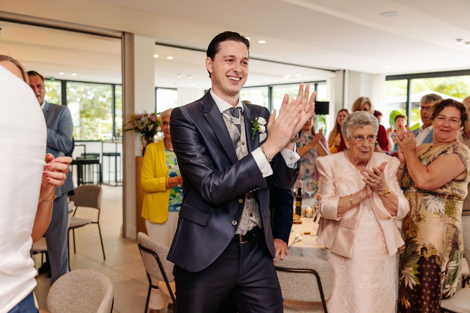 Smiling groom in navy suit claps during wedding reception surrounded by applauding family guests.