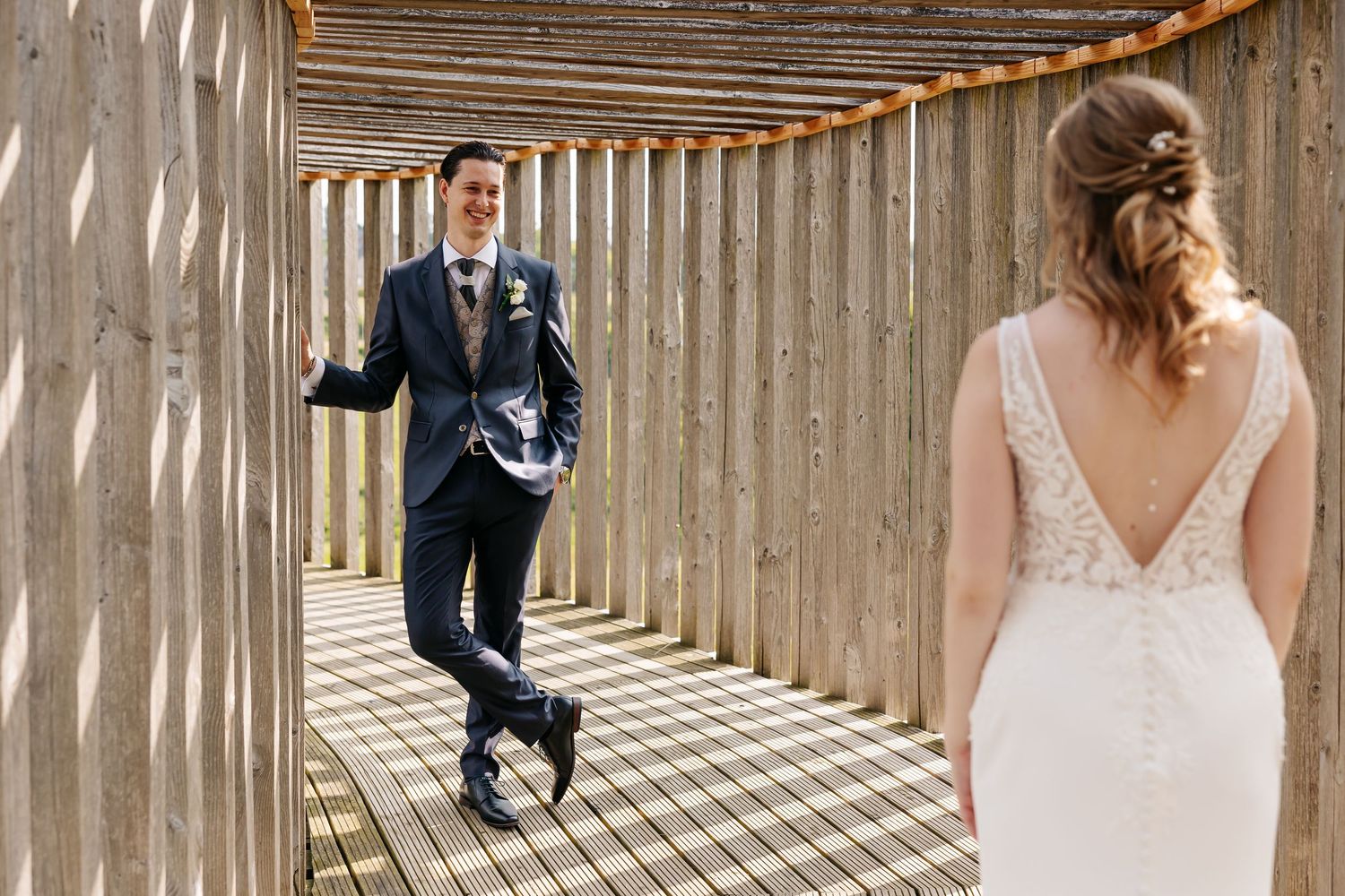 Groom in navy suit smiling at bride in lace wedding dress in wooden corridor.