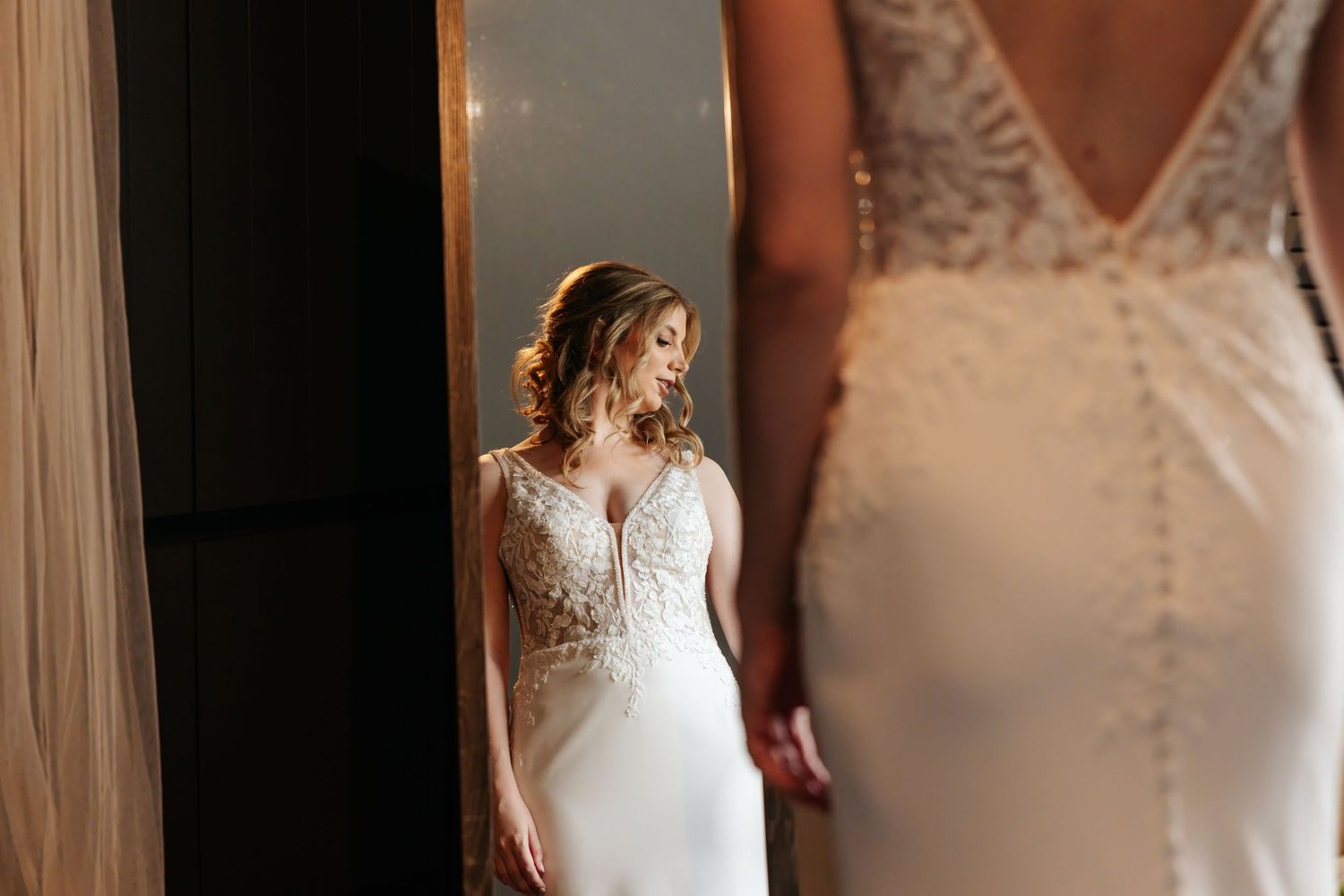 Bride in a fitted lace wedding dress admiring her reflection in a full-length gold-framed mirror.