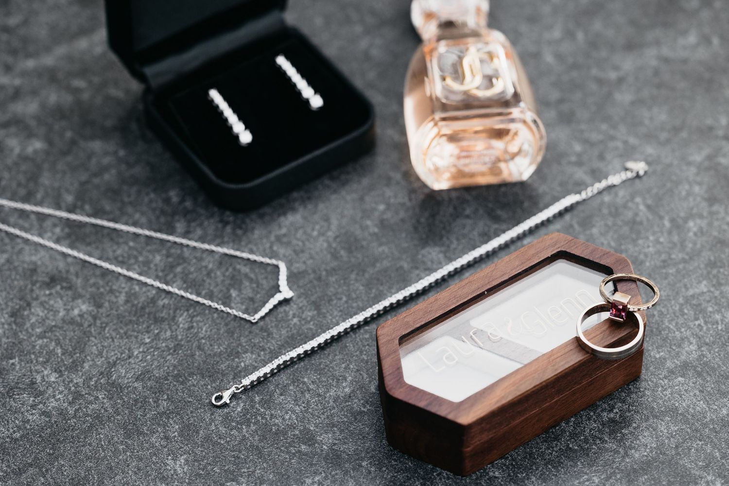 Bridal jewelry flat lay featuring diamond earrings, silver necklace, rings in wooden box, and perfume bottle.