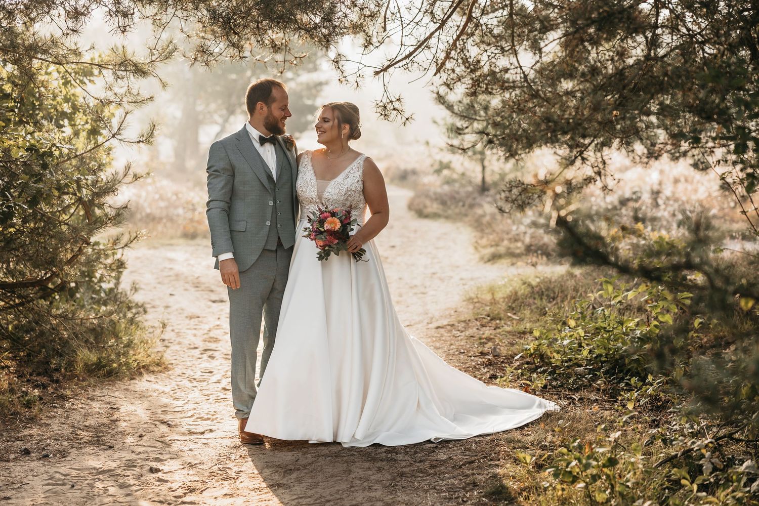 Bride and groom walk hand in hand on a sandy path through a sunlit forest in wedding attire.