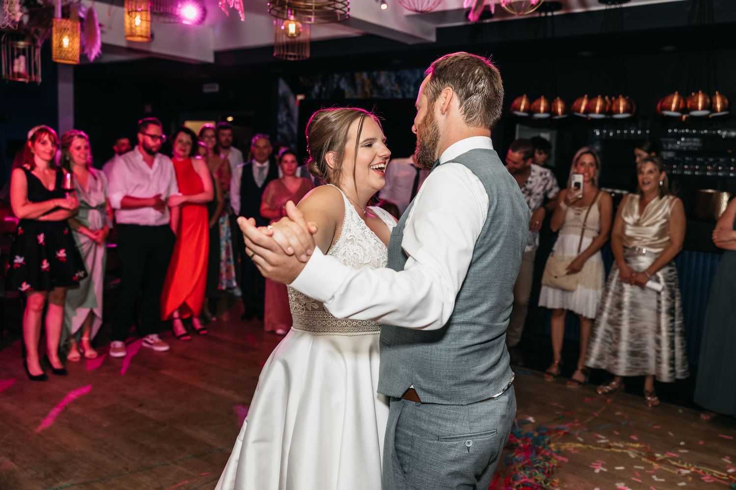 Bride and groom share their first dance while smiling guests clap and watch in a warmly lit wedding reception venue.