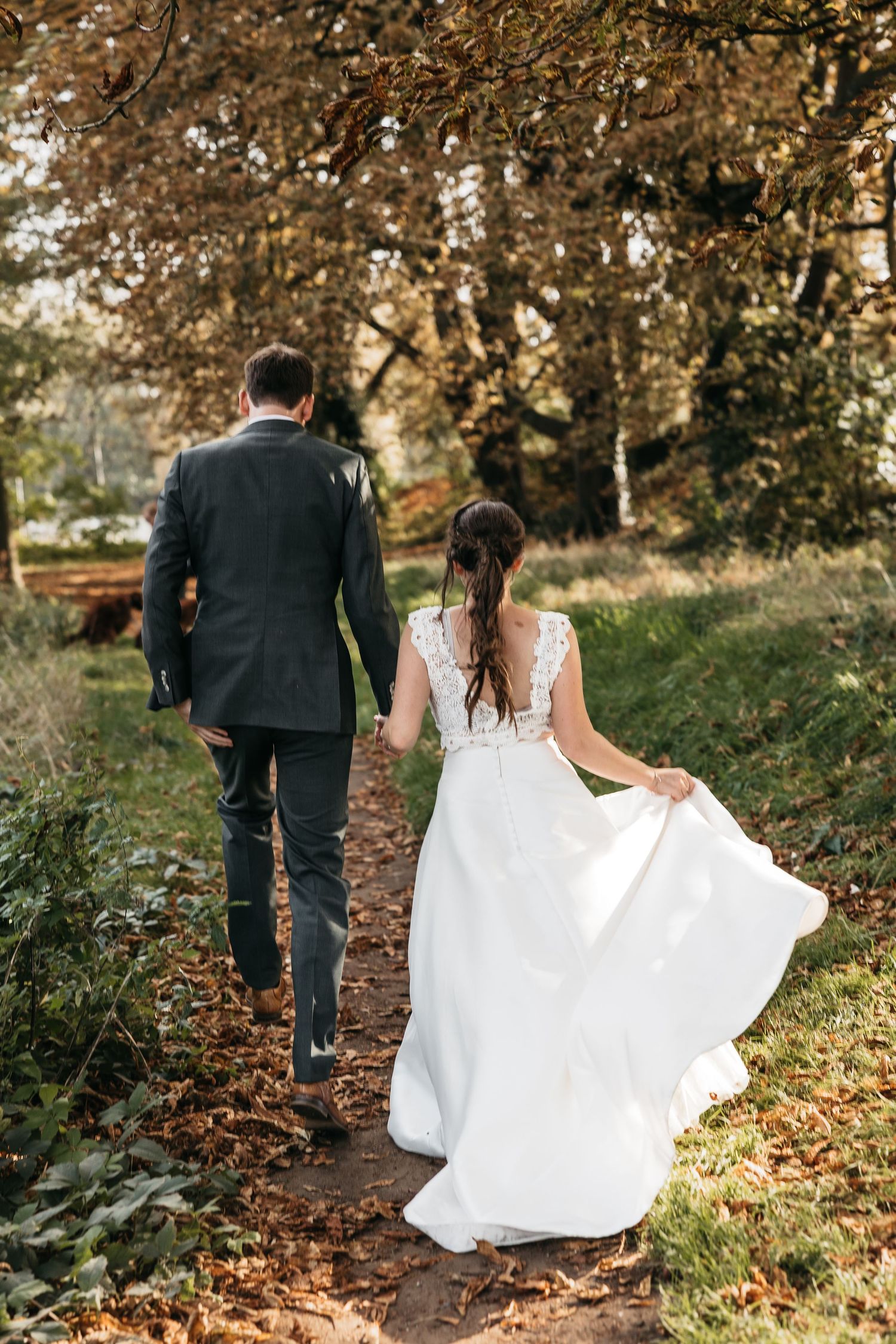 Bride in lace wedding gown and groom walking hand in hand down an autumn forest path.