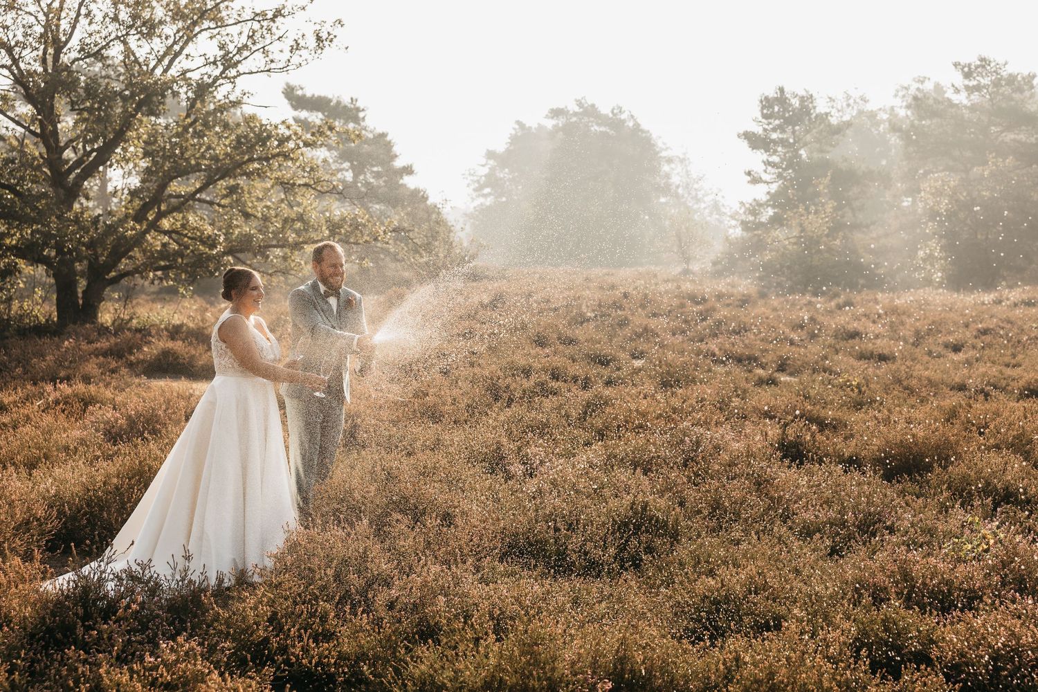 Newlyweds celebrating with champagne spray in a sunlit heather meadow on their wedding day.