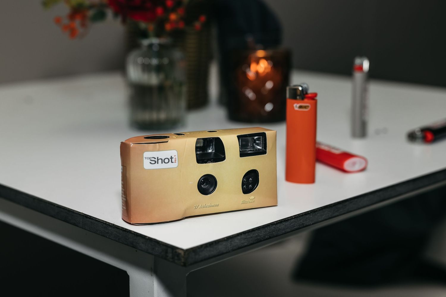 Disposable 1Shot film camera on white table with lighter and vase.