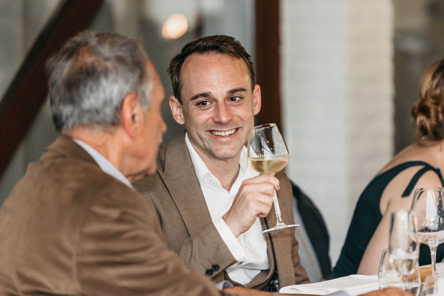 Smiling man raising a glass of white wine in a toast at an elegant dinner event.