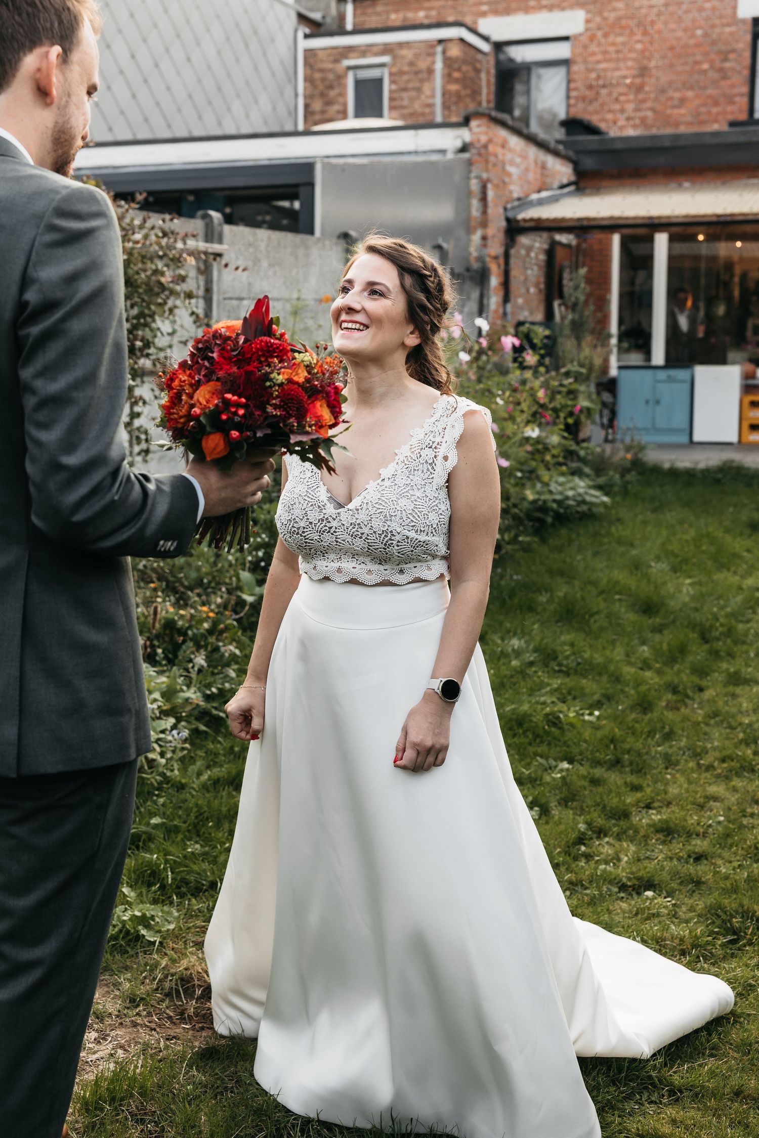 Bride in beaded white gown smiling as groom presents her with a rich red and orange bouquet.