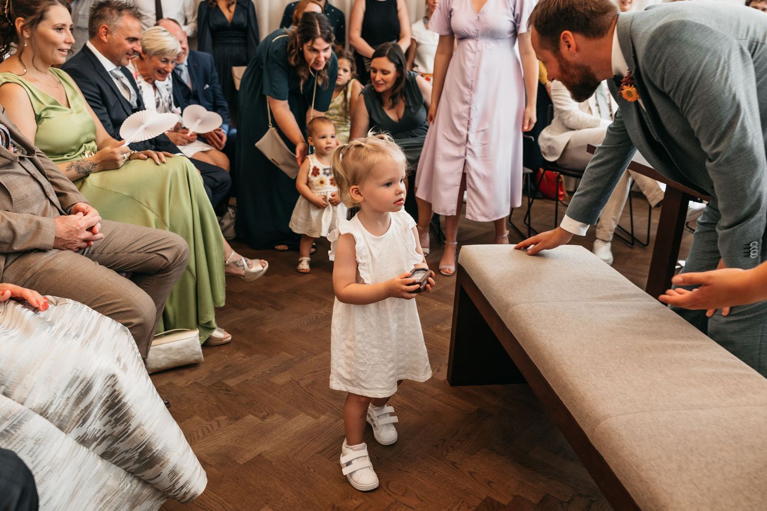 Adorable toddler girl in white dress laughs joyfully during a wedding ceremony surrounded by seated guests.
