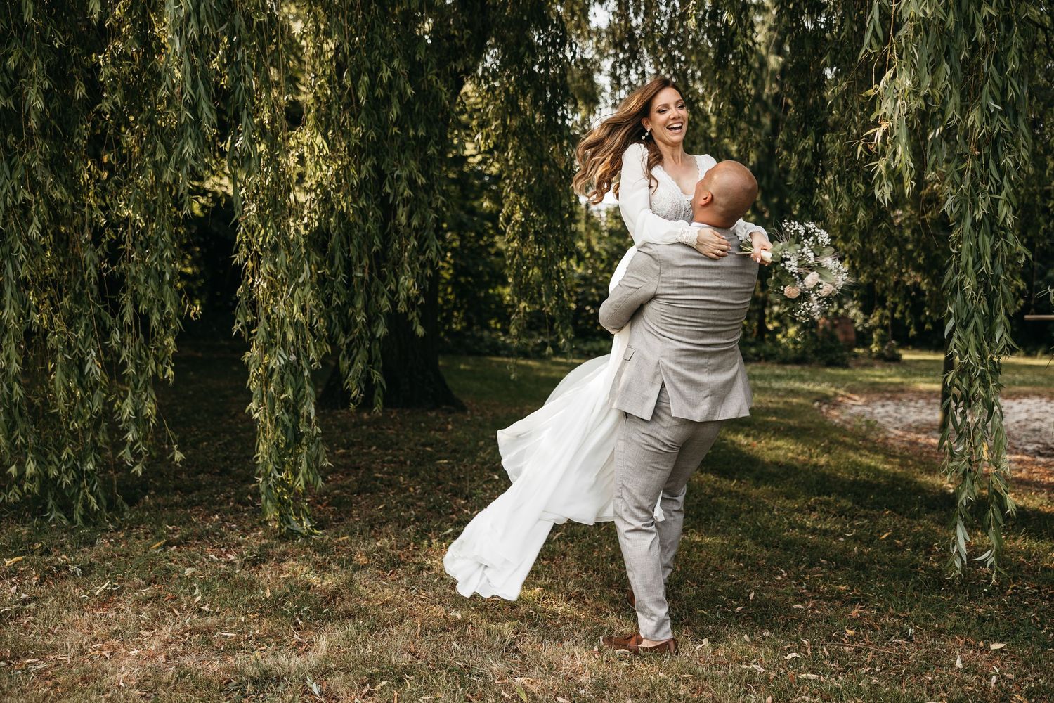 Groom lifting and spinning his laughing bride beneath weeping willow trees in a lush garden.