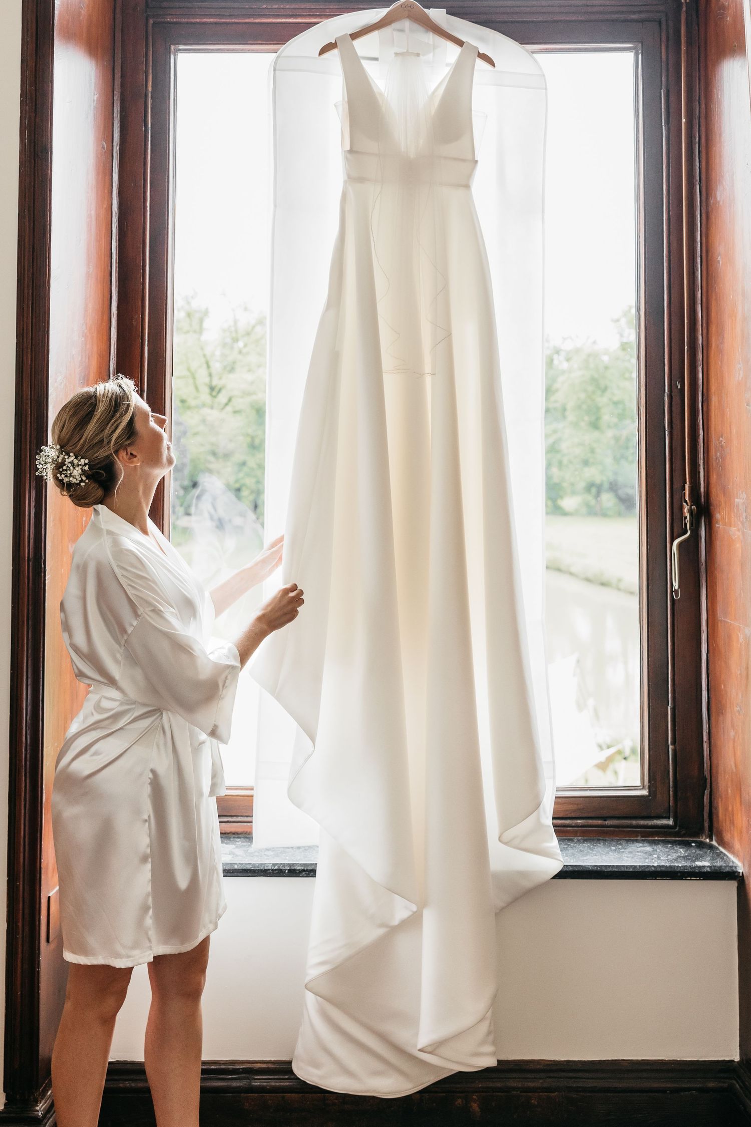 Bride in white robe admires elegant A-line wedding gown hanging in a sunlit window.