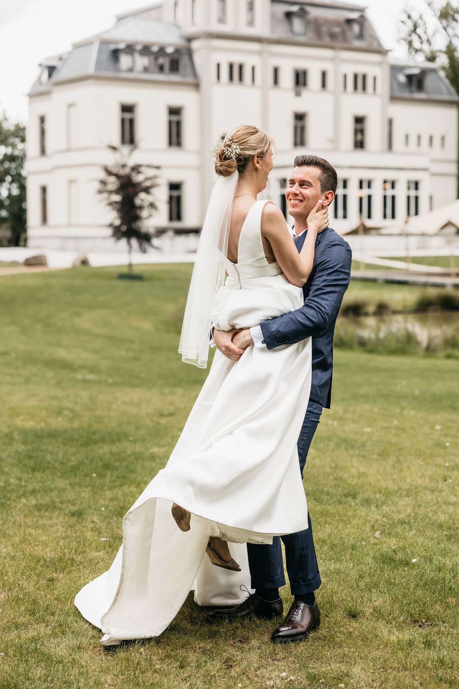 Bride and groom dancing joyfully on green lawn in front of elegant white manor house.
