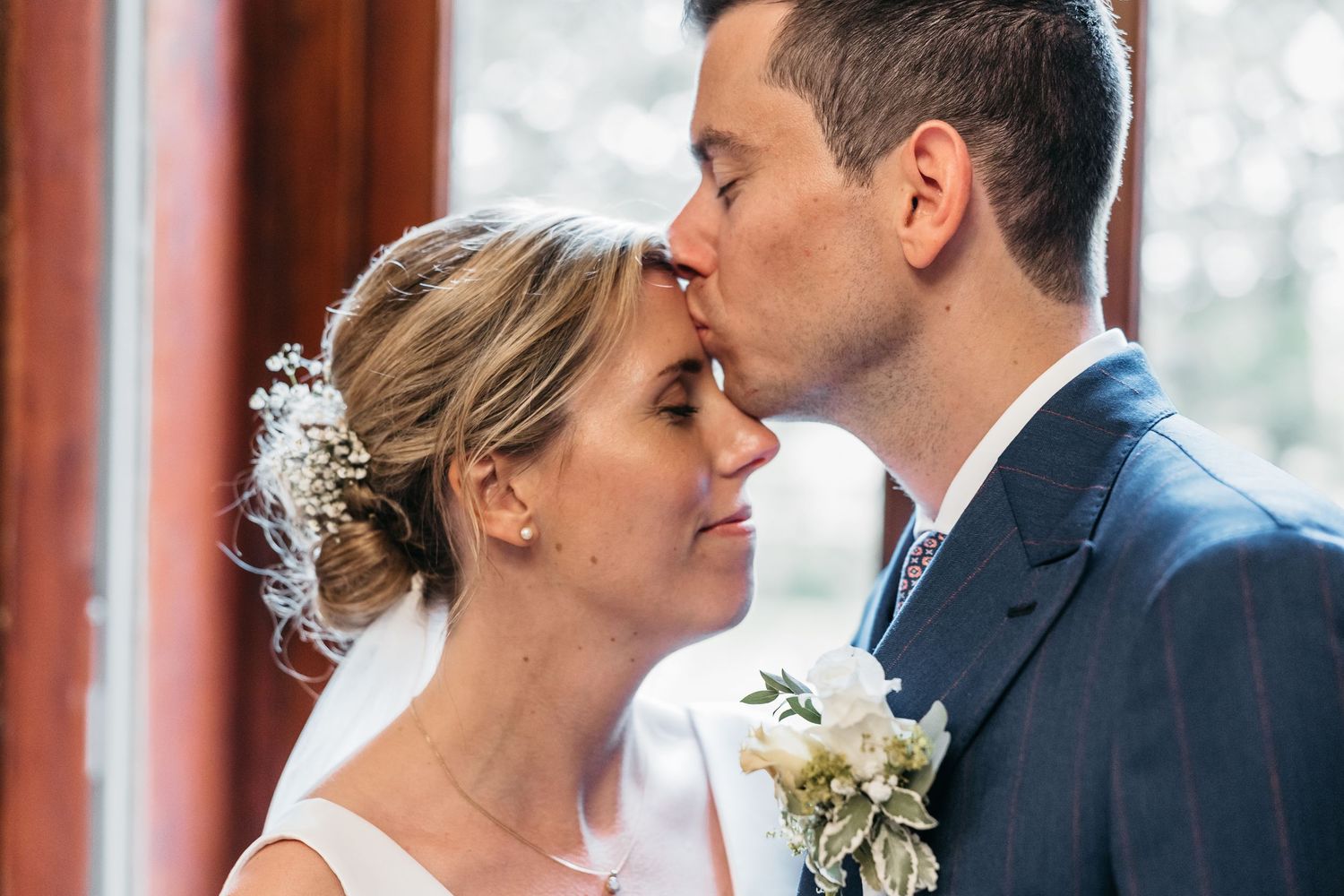 Groom tenderly kissing bride on forehead near window, bride wearing floral hair accessory and veil.